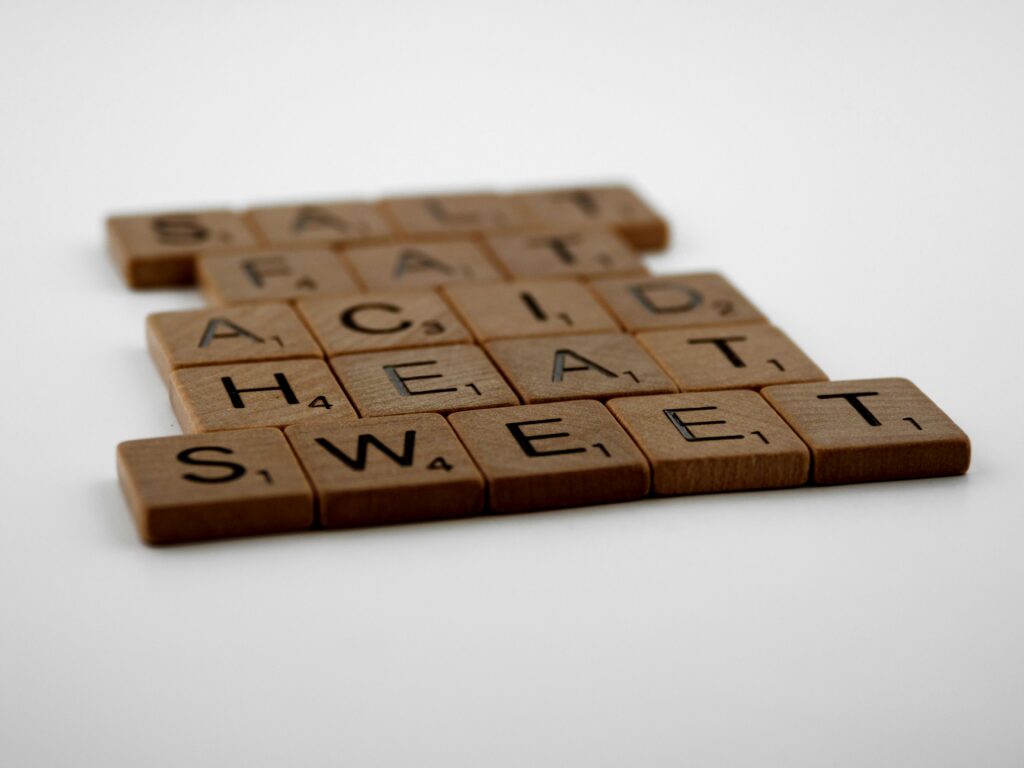 Wooden Scrabble tiles arranged to spell 'Salt, Fat, Acid, Heat, Sweet' on a white background.